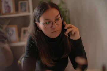 Focused young woman with glasses talking on the phone while working from home, sitting at a desk with framed photos and plants in the soft-lit background.