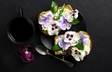 Breakfast cream cheese toasts with pansies culinary flowers on black plate, on black background, top view.