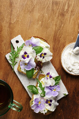 Breakfast cream cheese toasts with pansies flowers on ceramic board, on wooden background. top view, copy space