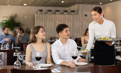 Young woman waitress brings order from couple young man and young woman in restaurant
