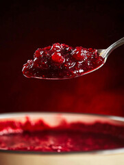 A photorealistic close-up of thick raspberry jam resting on a silver spoon held above a cream-colored enamel pot.