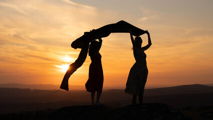 two women in silhouette hold a floating fabric or an abstract fourth figure against the vibrant...