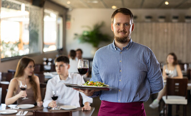 Professional friendly waiter holding serving tray with glass of red wine for restaurant guests