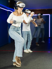In darkened hall of gaming club, girl in VR helmet takes part in group game. Player overcomes...