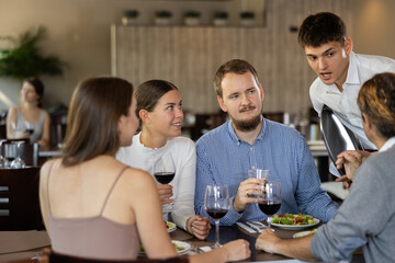 Young man waiter takes order from couples of men and women in restaurant