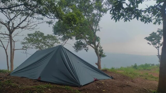 A dark-colored flysheet tent stands on a misty mountain peak, surrounded by tall trees and foggy skies, creating a serene and secluded camping atmosphere.