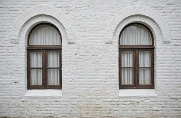 Fototapeta premium Two arched windows with wooden frames set in white brick wall of 18th-century historical building. Windows feature sheer white curtains, creating symmetrical facade pattern. Textured wall offers