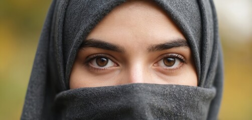 Naklejka premium Close-up portrait of beautiful Muslim woman eyes, partially veiled. Gaze direct, engaging, set against softly blurred outdoor background with warm, autumnal colors. Image emphasizes expressive eyes,
