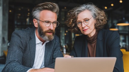 Mature team of partners sitting at table, business man and business woman discussing project on laptop in office. Two colleagues of professional business people working together.