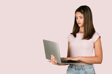 Teenage Caucasian brunette girl wearing a light pink shirt and jeans, holding and typing on a laptop, isolated on pastel pink background. Suitable for concepts related to technology, remote work