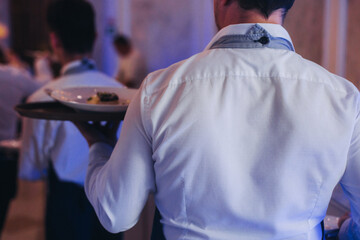 Waiter in uniform serving dishes on party event, server carrying plates with food at the table,...