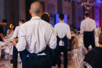 Waiter in uniform serving dishes on party event, server carrying plates with food at the table,...