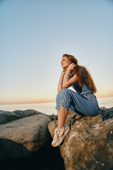 lifestyle woman in modern boho-western denim street style sits thoughtfully on rocks by the sea,...