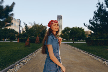 young woman wearing denim dress and red bandana walking in urban park during golden hour with...