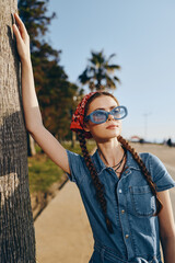 young woman in denim dress and blue sunglasses leaning on palm tree outdoors on sunny day with red...