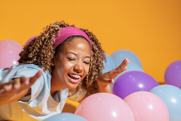 Young woman lying down having fun among colorful balloons on yellow background