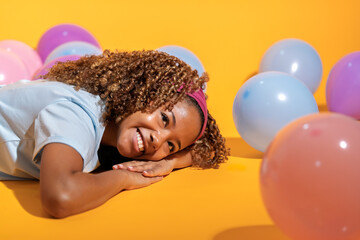 Young girl lying down and having fun surrounded by balloons on yellow background