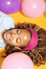 Young woman relaxing among colorful balloons on yellow background