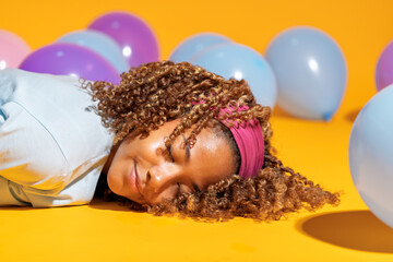 Young woman relaxing among colorful balloons on yellow background