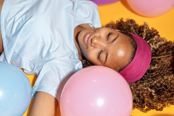 Young woman relaxing among colorful balloons on yellow background
