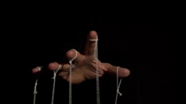 A hand manipulating puppet strings on black background