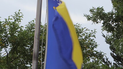 The national flag of the country of Bosnia and Hercegovina waving in a strong wind on top of a flag pole with green tress right behind it. 