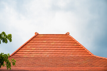 A terracotta tiled roof with decorative finials against a cloudy sky, partially obscured by green...