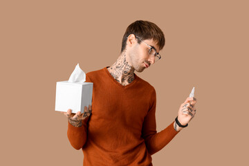 Allergic young man with nasal drops and tissues on brown background