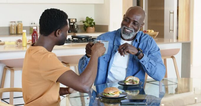 African American father and son at breakfast in kitchen with mugs reacting to noise, checking phone