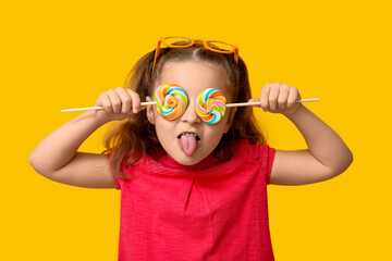 Cute little girl covering eyes with lollipops and showing tongue on color background