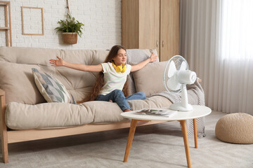 Young girl sitting on sofa with blowing electric fan in living room
