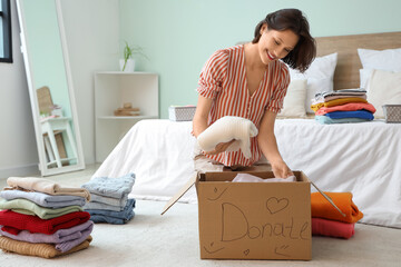Young pretty woman packing different clothes in donation cardboard box on floor at home