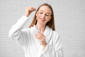 Beautiful young woman in bathrobe applying cosmetic oil on face in bathroom