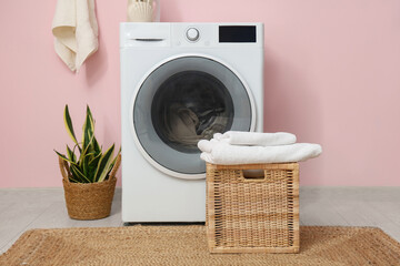 Modern washing machine with laundry, basket and houseplant near pink wall