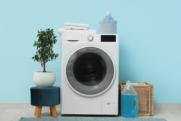 Modern washing machine with houseplant, basket and detergents near blue wall