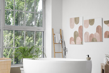 Stylish bathroom with bathtub, window and pictures on white wall