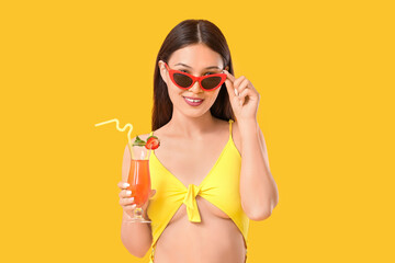 Beautiful young Asian woman in swimsuit and sunglasses with glass of cocktail on yellow background