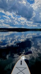 A stand-up paddleboarder’s view of a calm Nordic lake, perfectly reflecting the dramatic cumulus...