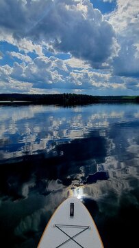A stand-up paddleboarder&rsquo;s view of a calm Nordic lake, perfectly reflecting the dramatic cumulus clouds above. The scene evokes serenity, freedom, and the intimate connection with nature, captured fro