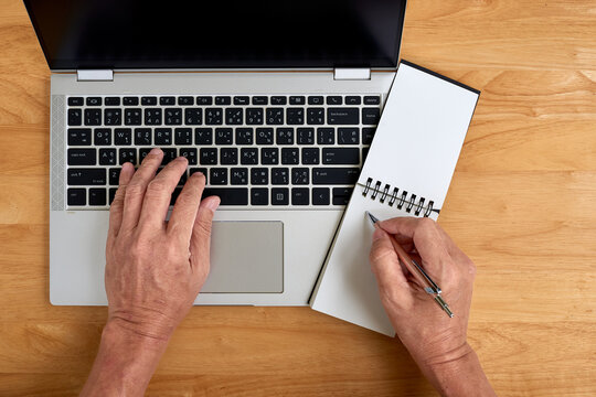 Top view of senior man's hands typing on Thai keyboard laptop and writing notes.
