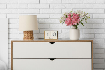 Cube calendar with date 10 June, lamp and flowers on chest of drawers near light brick wall