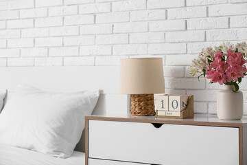 Cube calendar, lamp and beautiful flowers on chest of drawers near bed