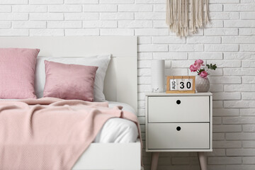 Bedside table with calendar, flowers, lamp and comfortable bed near light brick wall