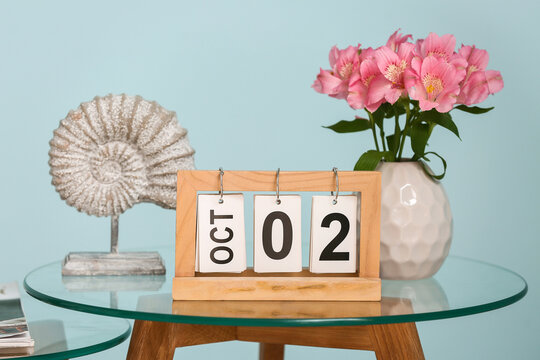 Wooden calendar with date 2 October and vase of beautiful flowers on coffee table near blue wall in living room