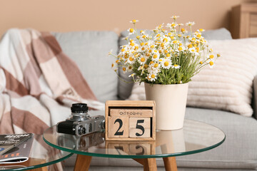 Cube calendar with date 25 OCTOBER, chamomiles and photo camera on table in living room
