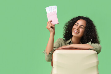 Young female African-American tourist with passport, tickets and suitcase on green background
