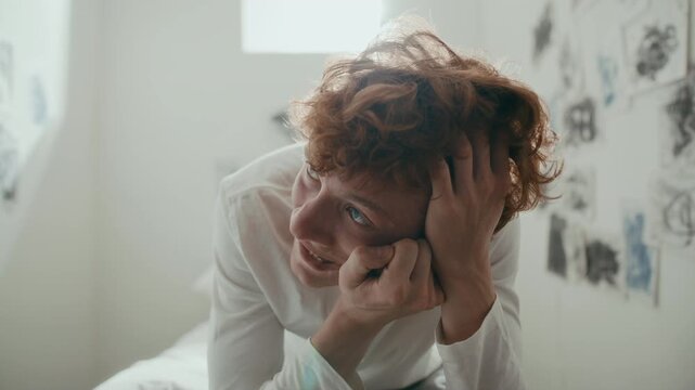 Young distressed male patient gripping his head and contorting face as experiencing psychotic episode while sitting on bed in sunlit hospital ward. Zoom-in shot