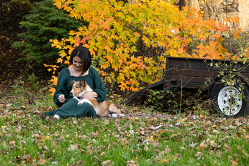young woman sitting on the grass with a sheltie puppy on her lap with a colorful fall foliage in...