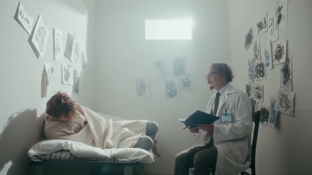 Doctor walking into psychiatric isolation room, sitting on chair, reading notes and talking to young male patient restrained in straightjacket on bed
