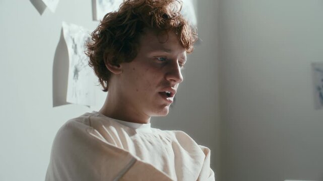 Young male patient in straitjacket sitting in psychiatric hospital ward with abstract drawings on walls, telling about his mental health to doctor during therapy session